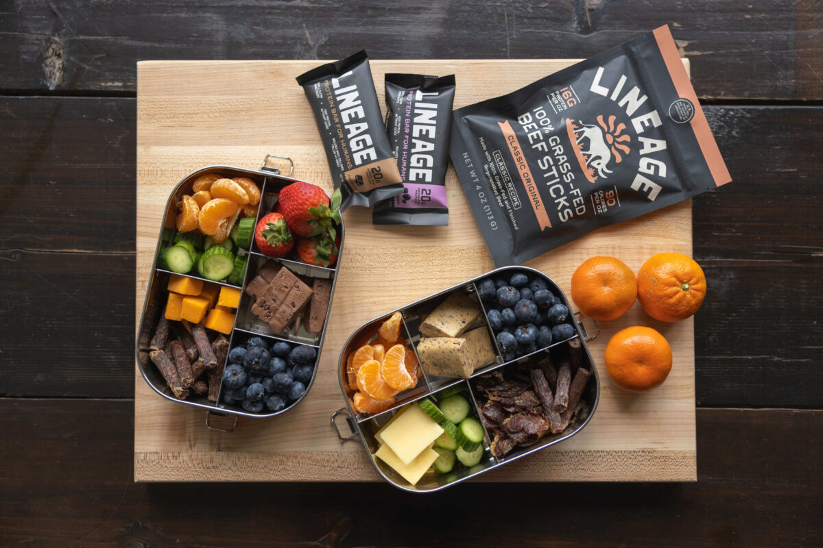 Lineage protein bars and beef sticks packed in lunch boxes with fruit, cheese and jerky